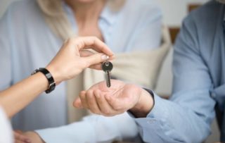 A person handing a key over to a couple, like how Windsor Pacific will hand you the key to one of our rental homes if you qualify.